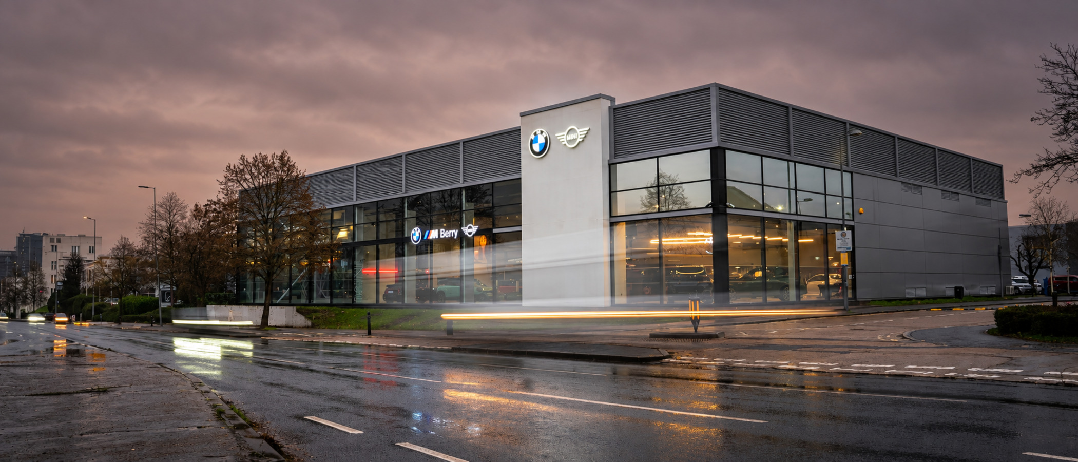 Berry's Chiswick BMW and MINI centre at dusk, a glass-fronted showroom with BMW and MINI signs lit against a wet road and passing headlights.