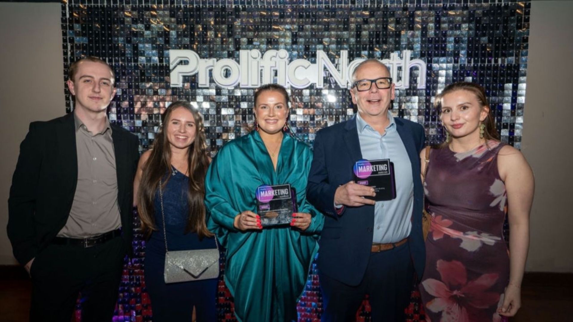 Five people at the Prolific North Marketing Awards in front of a sequinned Prolific North backdrop, two of them holding Marketing Awards winner trophies.