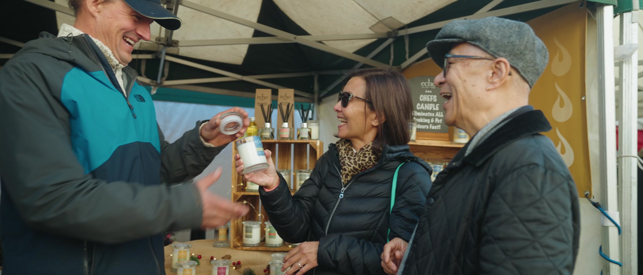 Three people laughing at a candle stall under a market canopy, where a stallholder in a cap lifts the lid from a candle jar while a woman in a leopard-print scarf holds it and an older man in a flat cap and glasses looks on