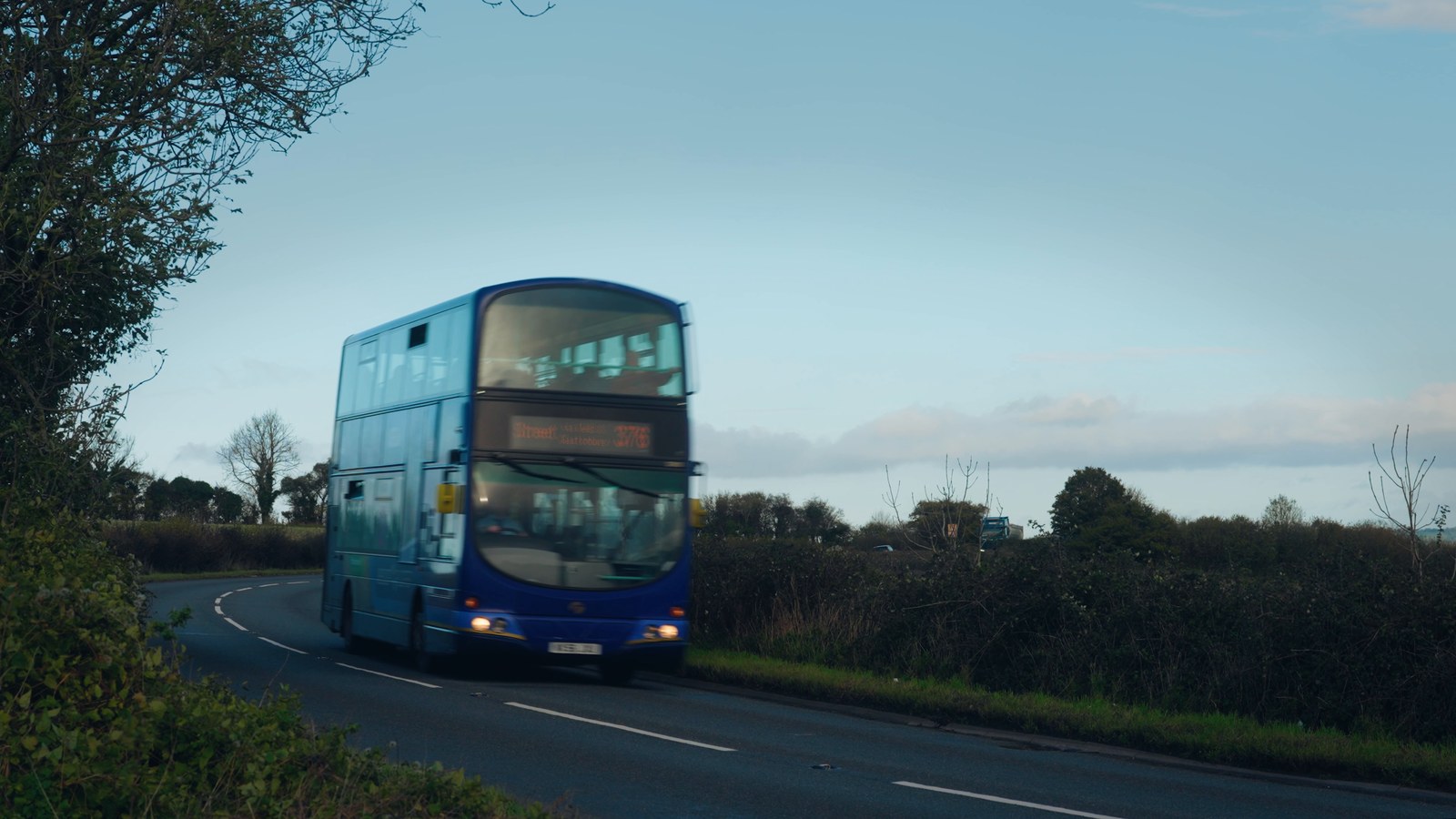 A First Bus double-decker with its headlights on, coming round a bend on a country road at dusk between hedgerows and bare trees