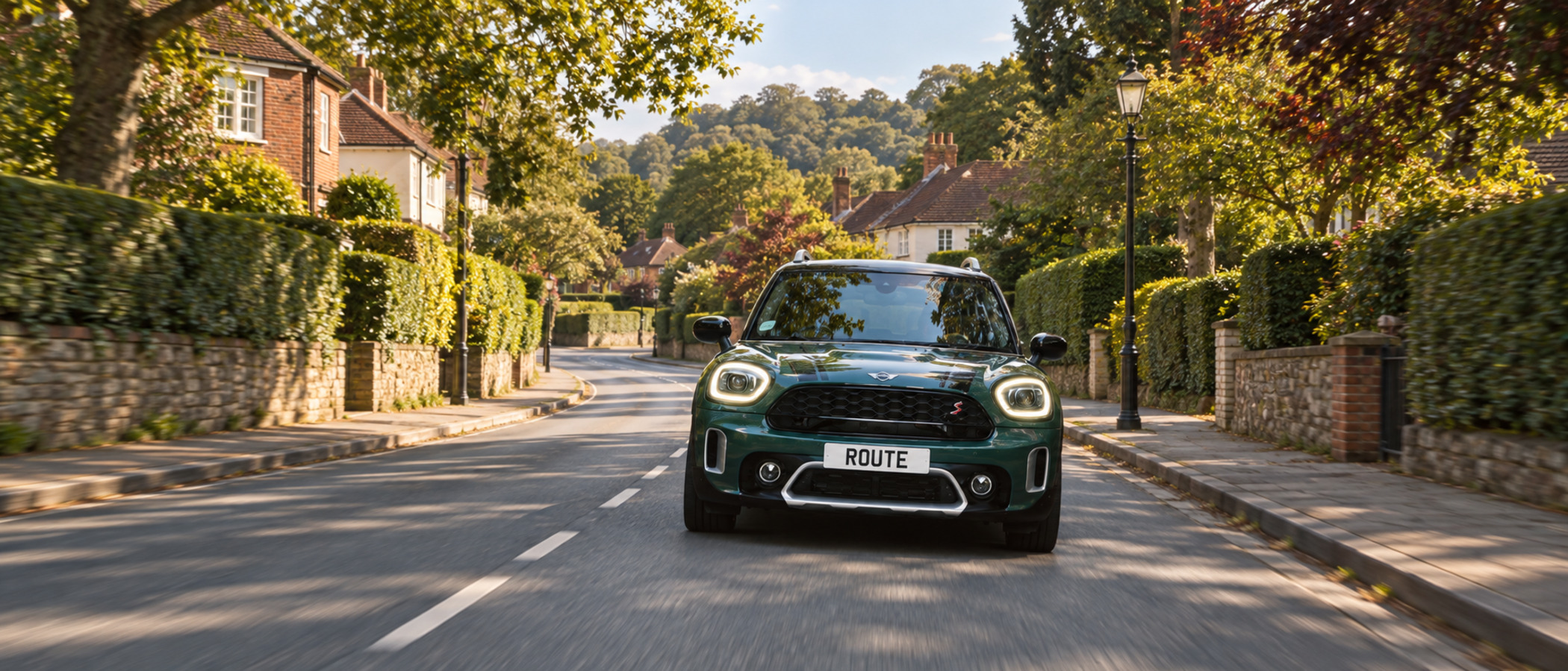 Illustrative image of a dark green MINI Countryman with its headlights on, driving along a sunlit tree-lined village street of brick houses, hedges and stone walls, carrying a ROUTE numberplate. Based on the campaign's video creative, not an original campaign asset.