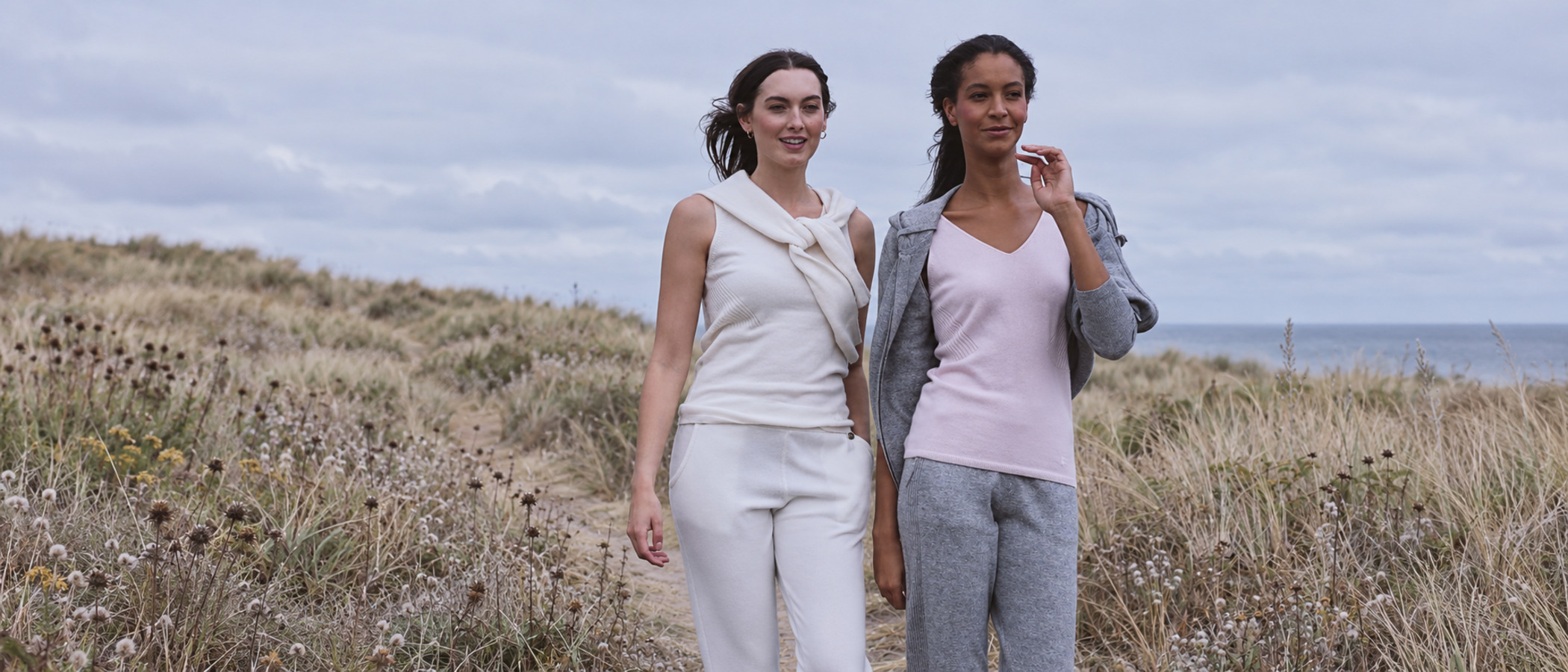 Two women in knitwear walking a path through coastal dunes under a grey sky, one in a cream sleeveless top and trousers, the other in a pale pink top and grey knitted joggers.