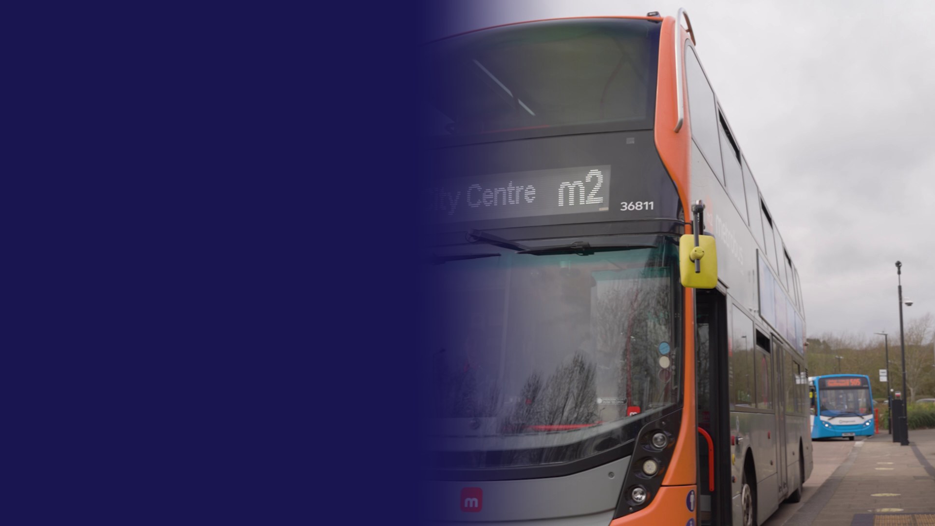 An m2 MetroBus double-decker in orange and grey showing City Centre m2 at a stop, with a blue bus waiting further along under a grey sky.