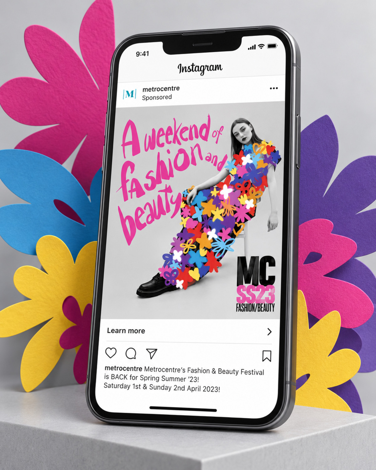 Illustrative layout of a Metrocentre Instagram ad on a phone, showing a model in a dress of paper flowers beneath the line A weekend of fashion and beauty, with the MC SS23 Fashion/Beauty mark and a Learn more button. Suggested copy, not an original campaign screenshot.