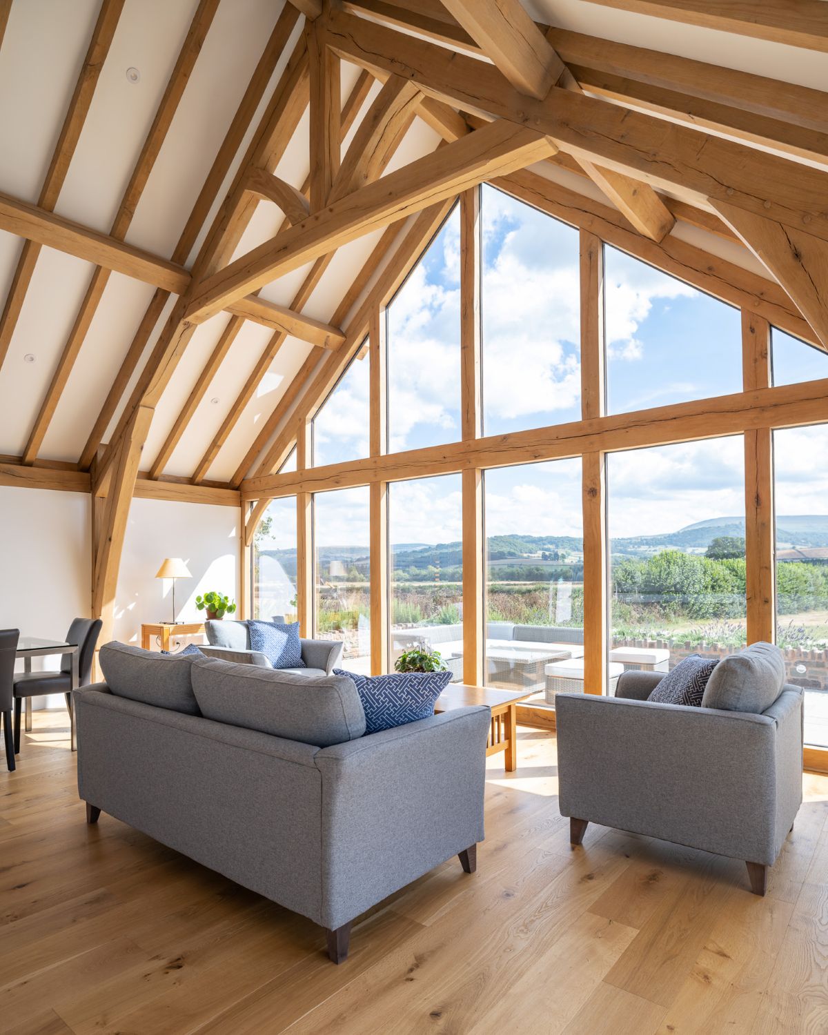 Vaulted oak-framed living room with a full-height glazed gable looking out over fields and hills, grey sofas on an oak floor
