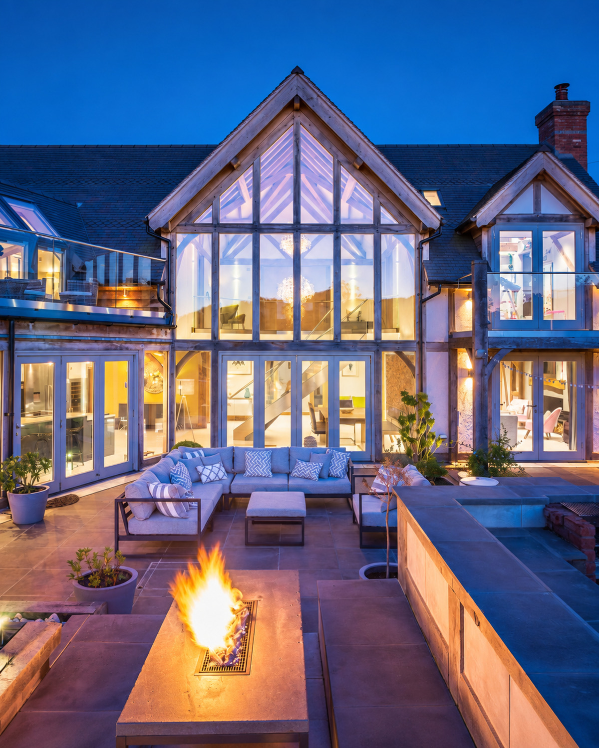Oak-framed self-build home at dusk, its full-height glazed gable lit from inside above a terrace with an outdoor sofa and a fire table